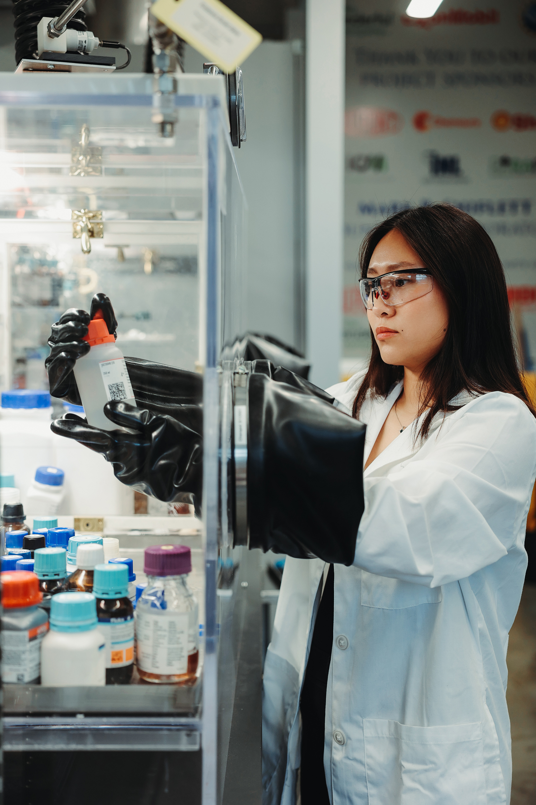 A researcher looks at a chemical bottle through a protective shield