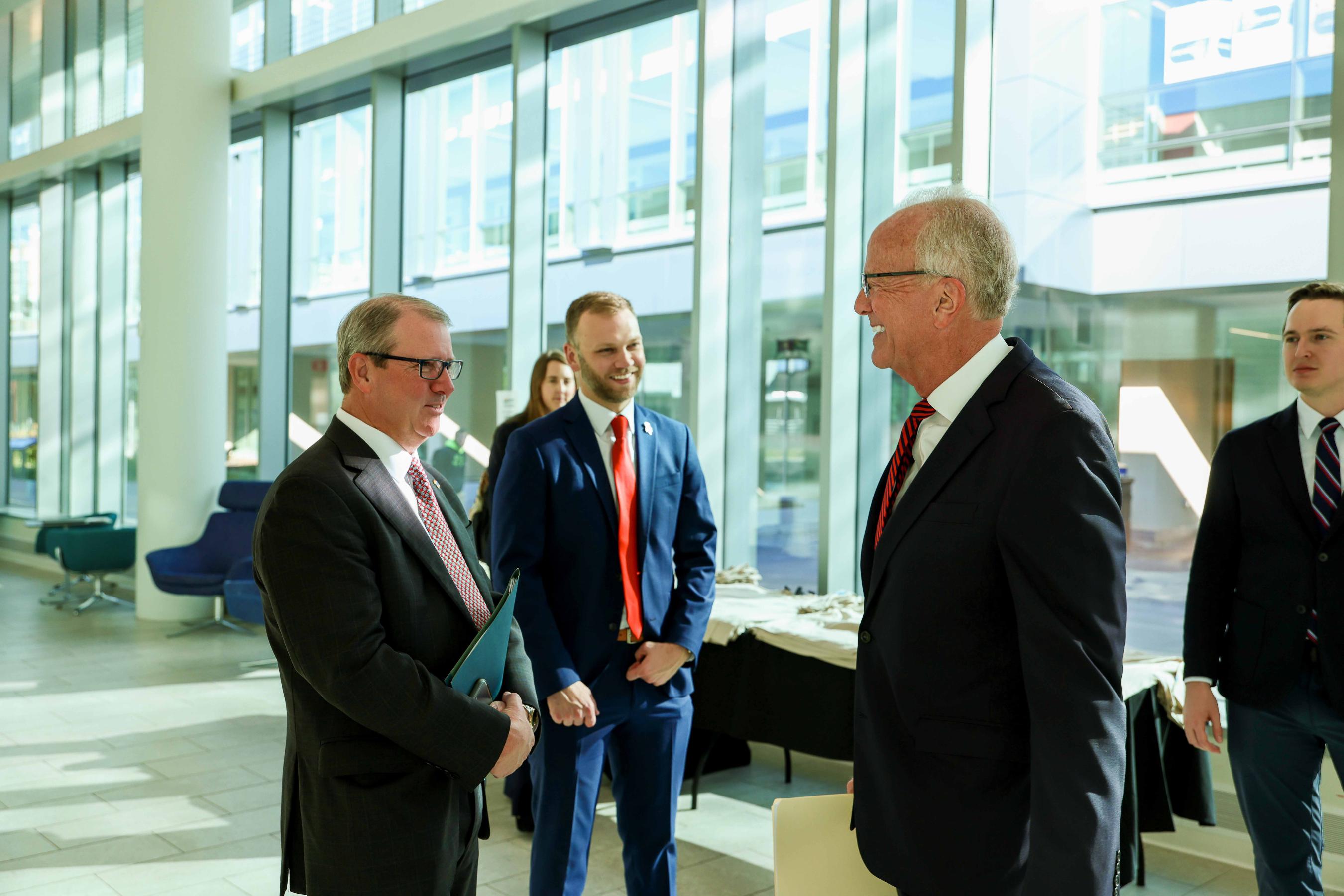 A group of people in business attire conversing in what appears to be a lobby or atrium with large windows.